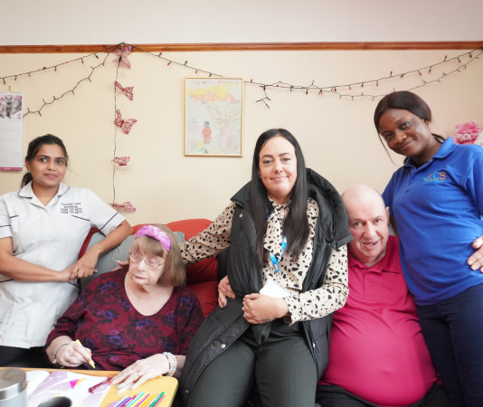 Group of caregivers and elderly individuals smiling together in a cozy room, representing compassionate community care, which is what we are about.