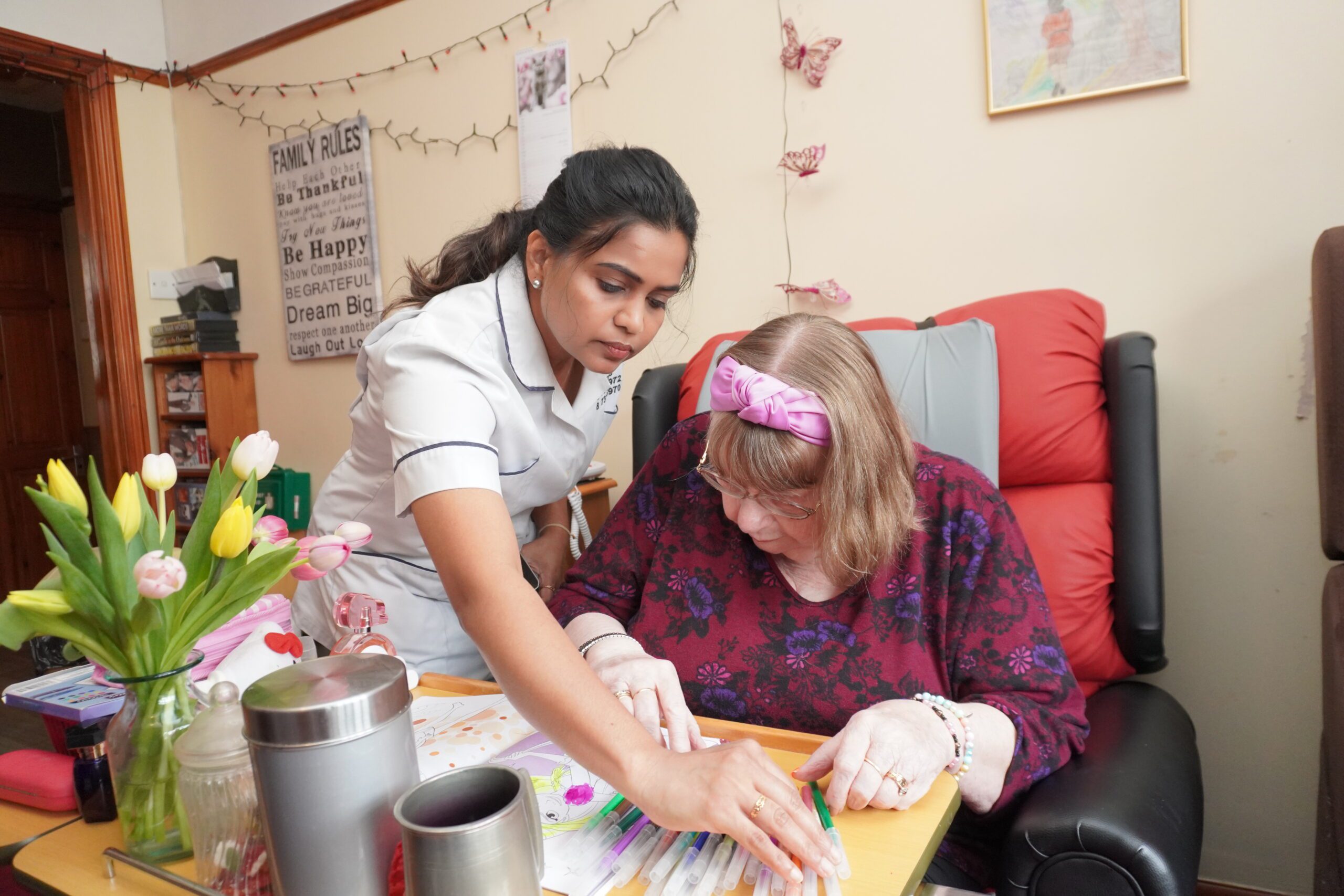 A caregiver assists an elderly woman with art supplies, providing guidance and support during an activity in a comfortable home environment.