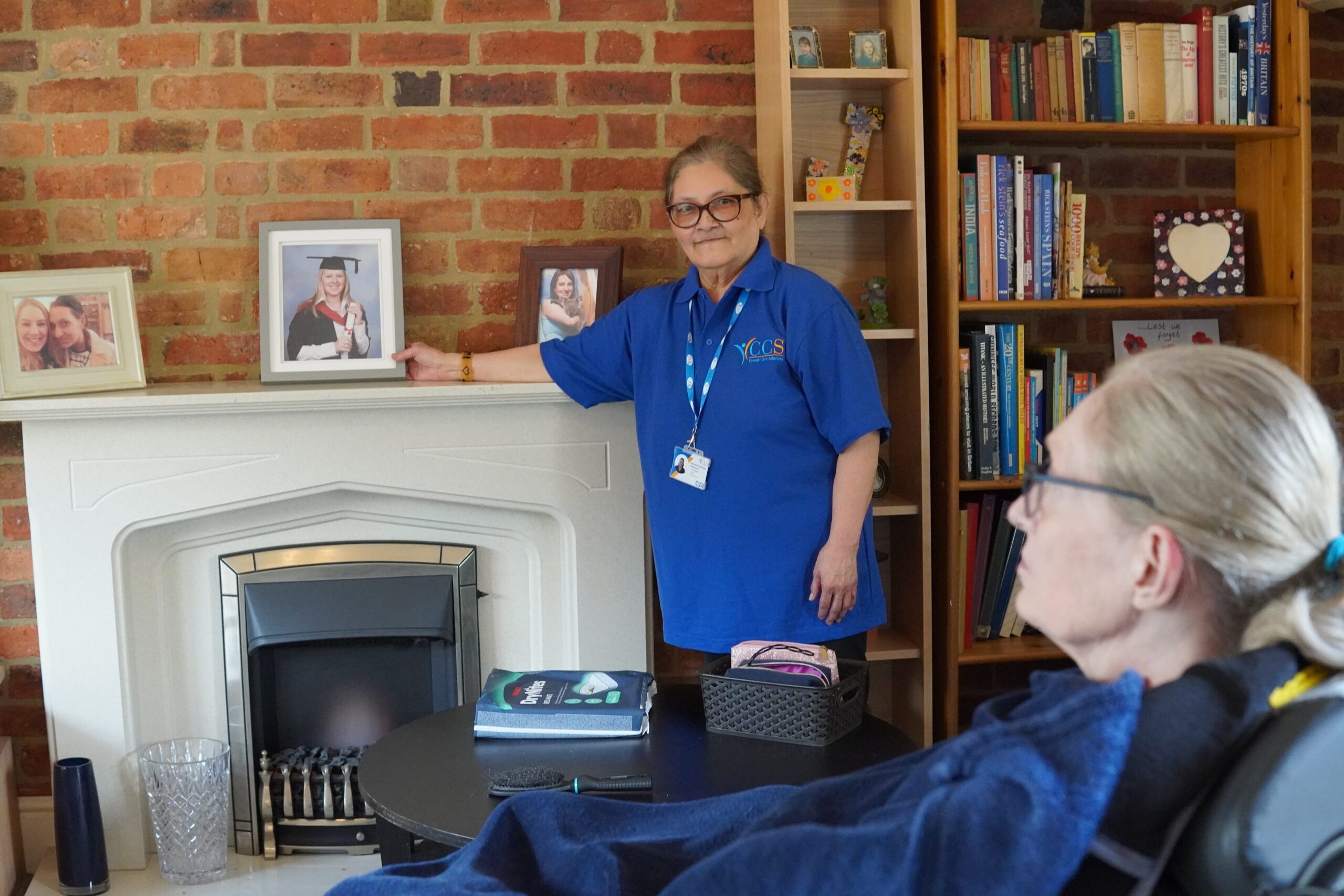 A caregiver from Concept Care Community Services, wearing a blue uniform, stands by a fireplace in a home setting, while a person in a wheelchair looks on, showcasing the personalised and attentive care provided.