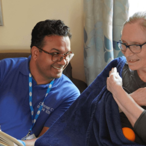 A caregiver from Concept Care Community Services sitting with an elderly woman, both smiling warmly while reading a book together.