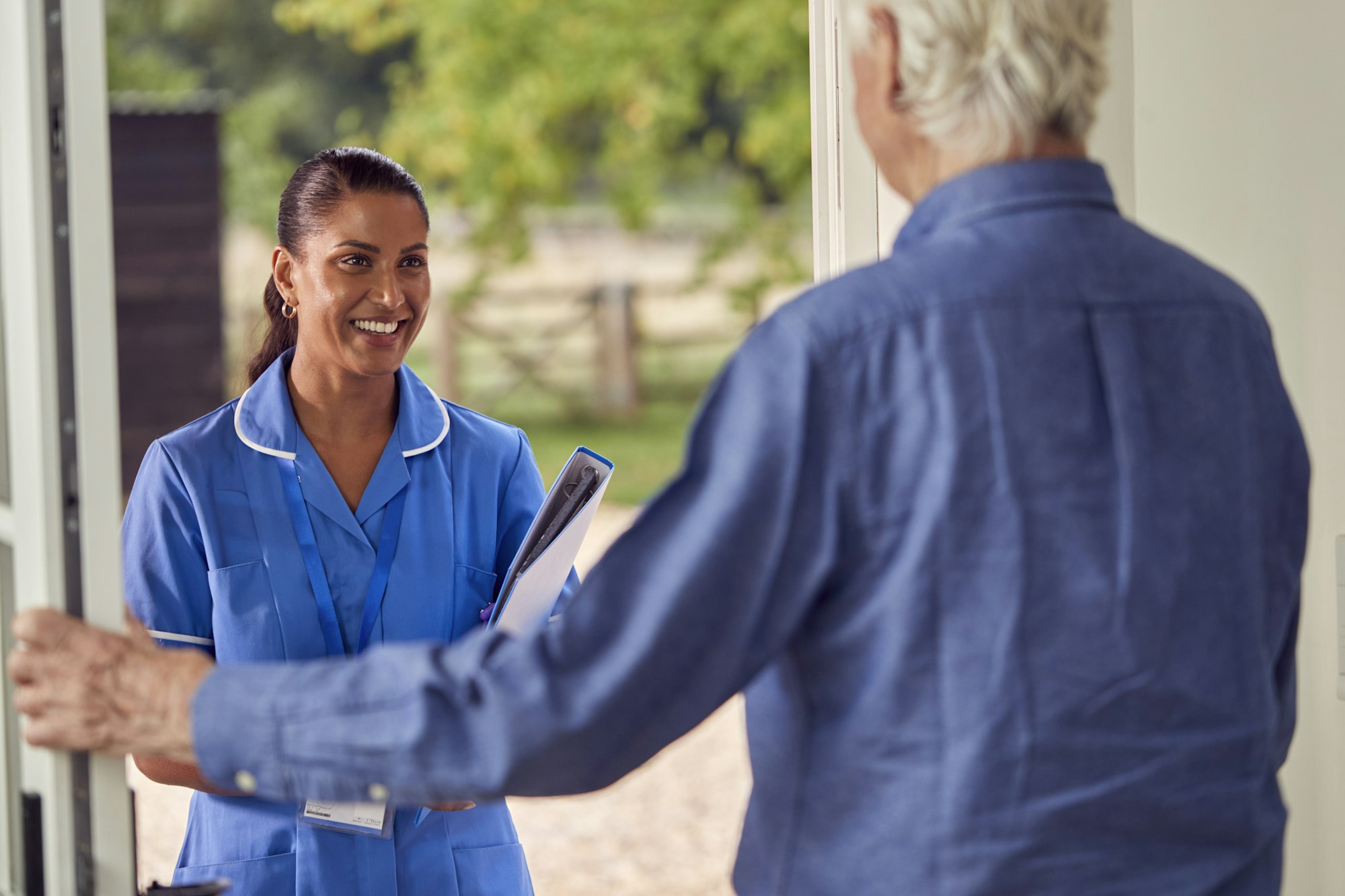 Senior Man Greeting Female Nurse Or Care Worker Making Home Visit In Uniform At Door