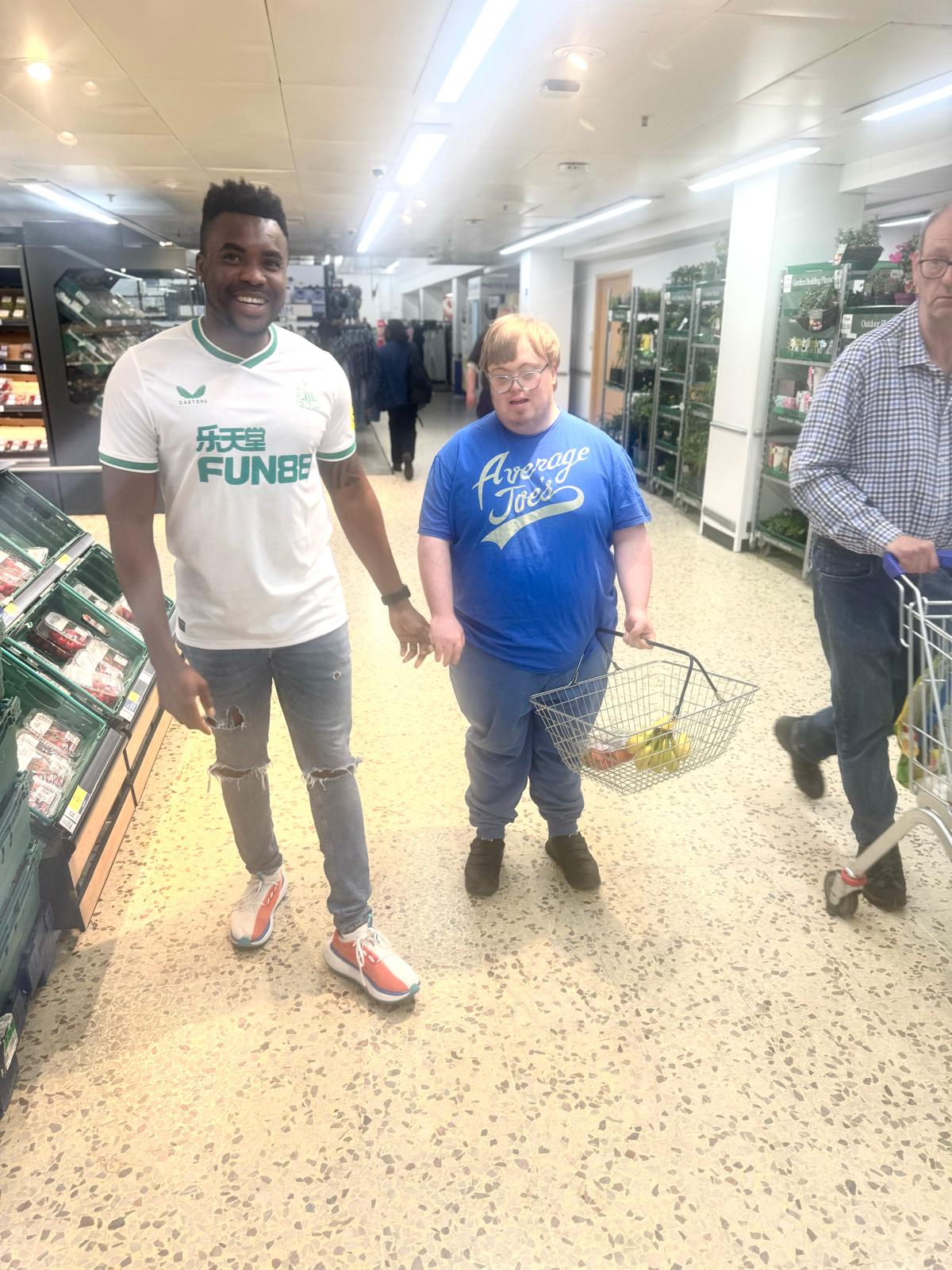 A carer supporting a client with their shopping in a supermarket aisle, both smiling and walking together.