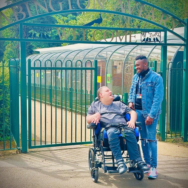 A carer and a client in a wheelchair smiling together outside the entrance of Abington Park, enjoying a day out.