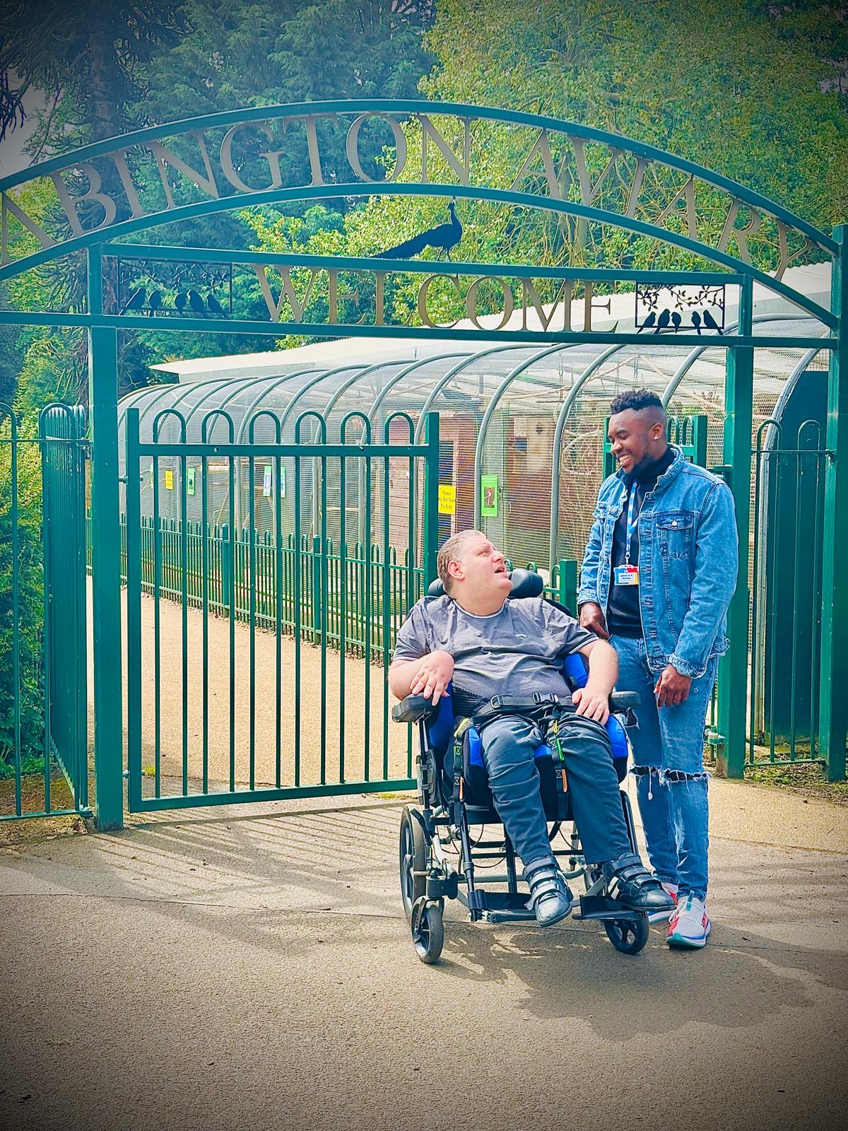 A carer and a client in a wheelchair smiling together outside the entrance of Abington Park, enjoying a day out.