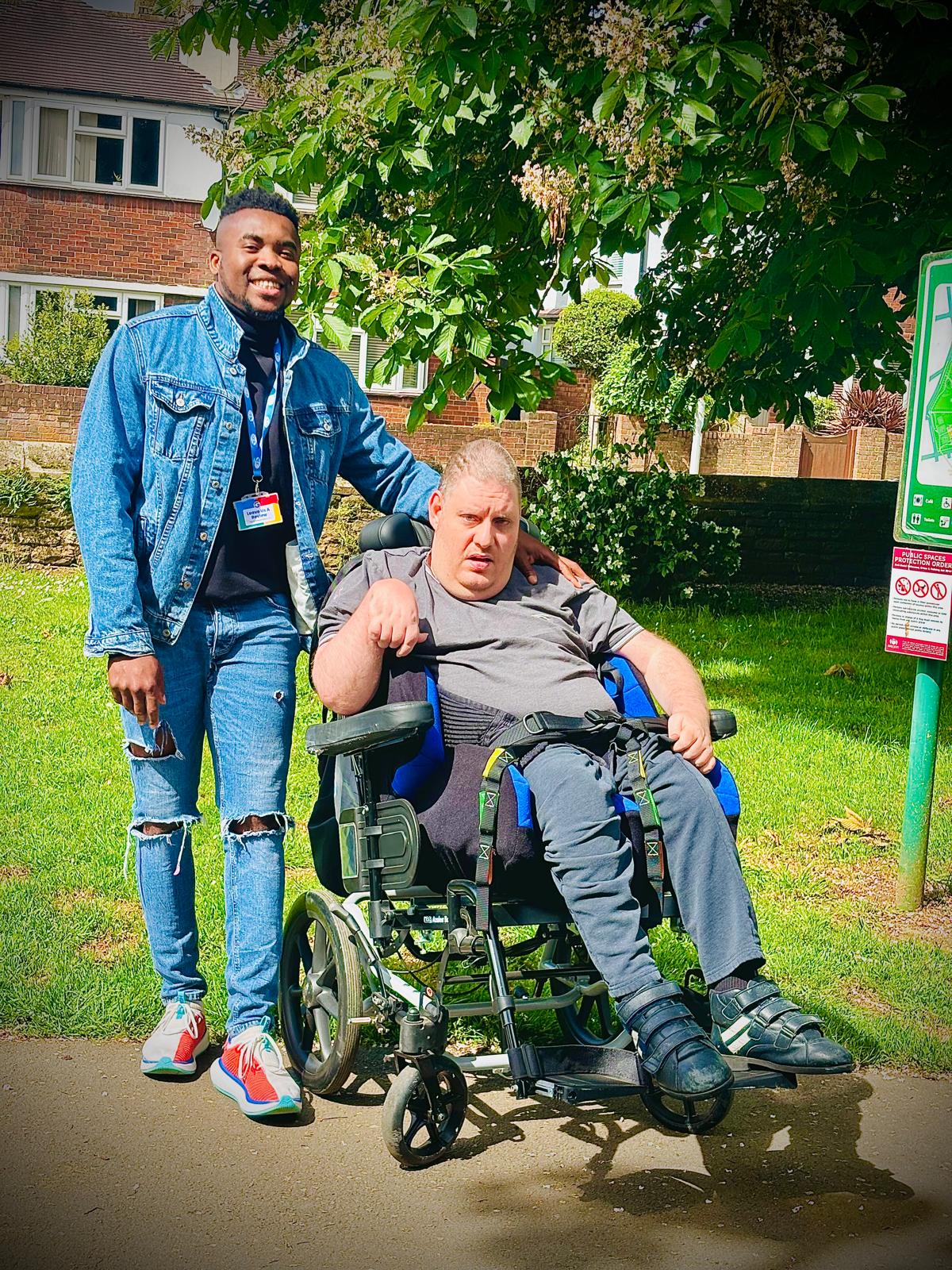 A carer smiling beside a client in a wheelchair as they enjoy a sunny day outdoors in a green park area.