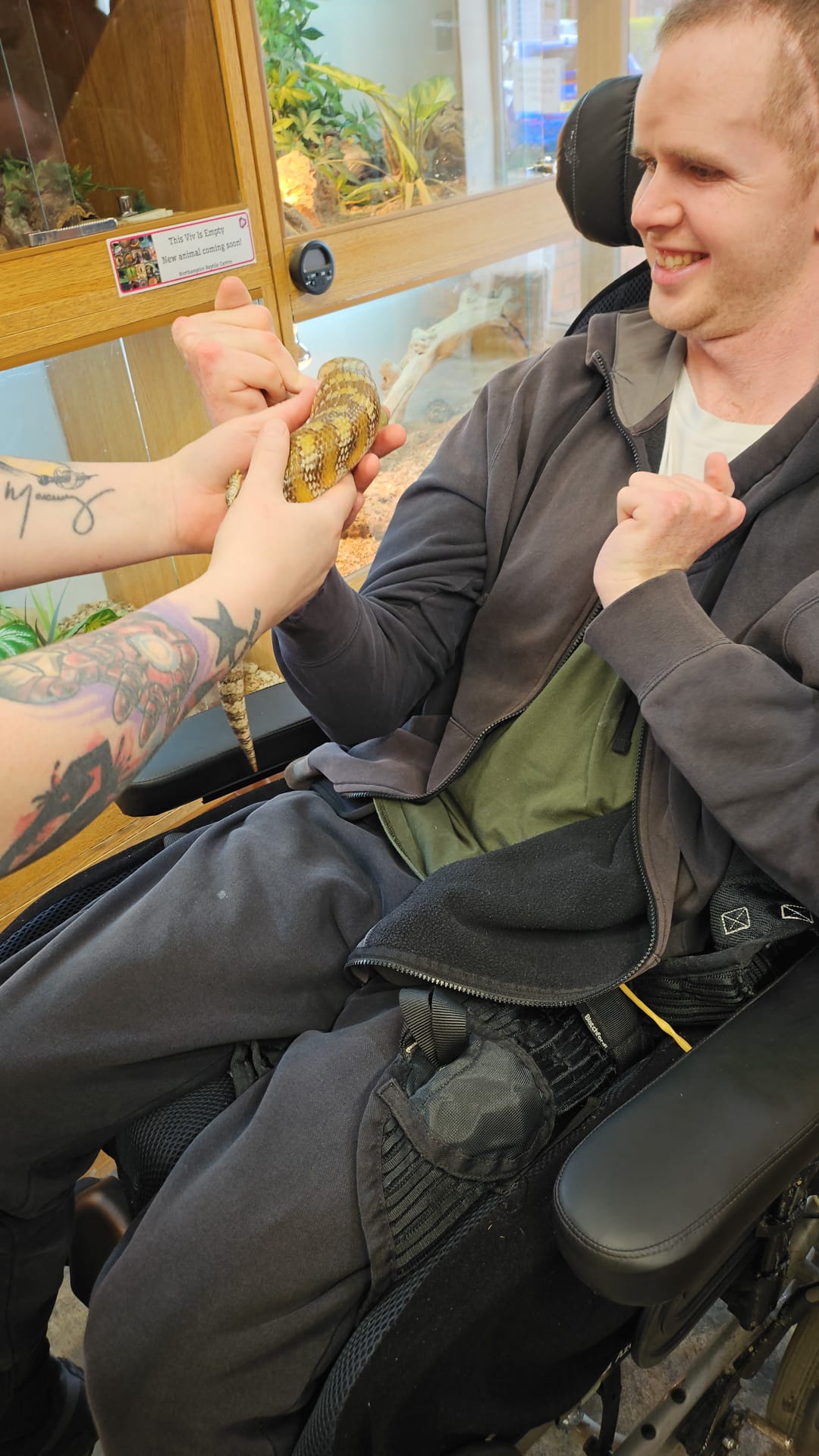 A smiling client in a wheelchair interacting with a reptile, held by a staff member at a zoo or animal centre.